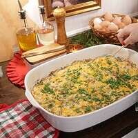 A woman in a kitchen spooning out a casserole from a 9x13" casserole white pan on wooden counter top.