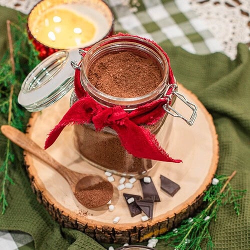 Overhead view of a glass jar of homemade hot cocoa mix tied with a red ribbon on a wood slice, with a wooden spoon of cocoa mix, dark chocolate pieces, and mini marshmallows, beside a glass mug of hot cocoa topped with marshmallows on a green gingham cloth