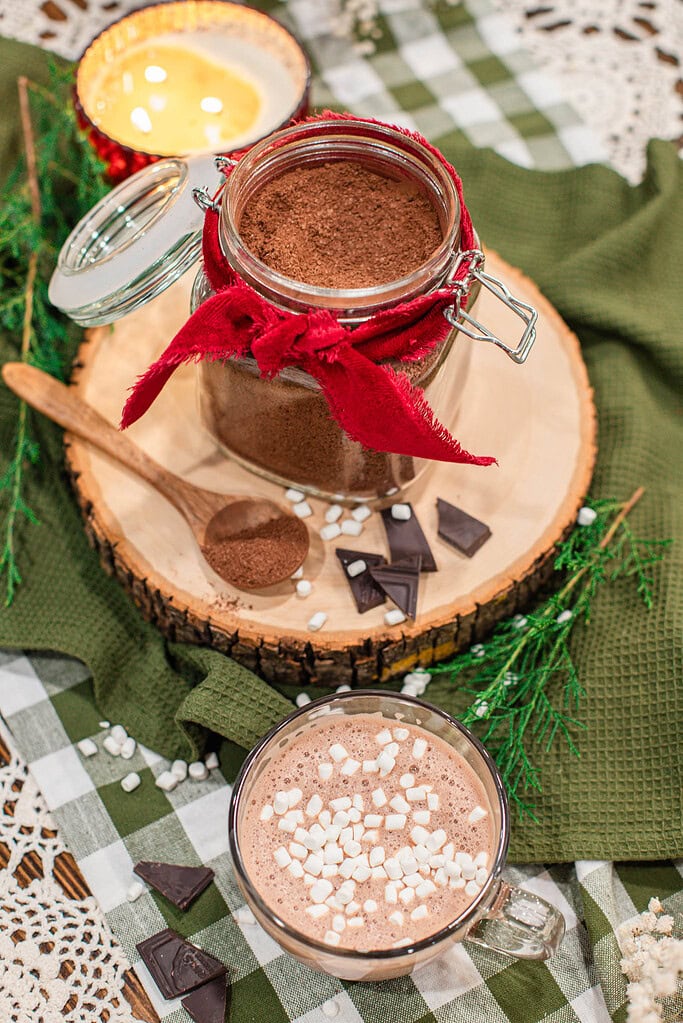 Overhead view of a glass jar of homemade hot cocoa mix tied with a red ribbon on a wood slice, with a wooden spoon of cocoa mix, dark chocolate pieces, and mini marshmallows, beside a glass mug of hot cocoa topped with marshmallows on a green gingham cloth