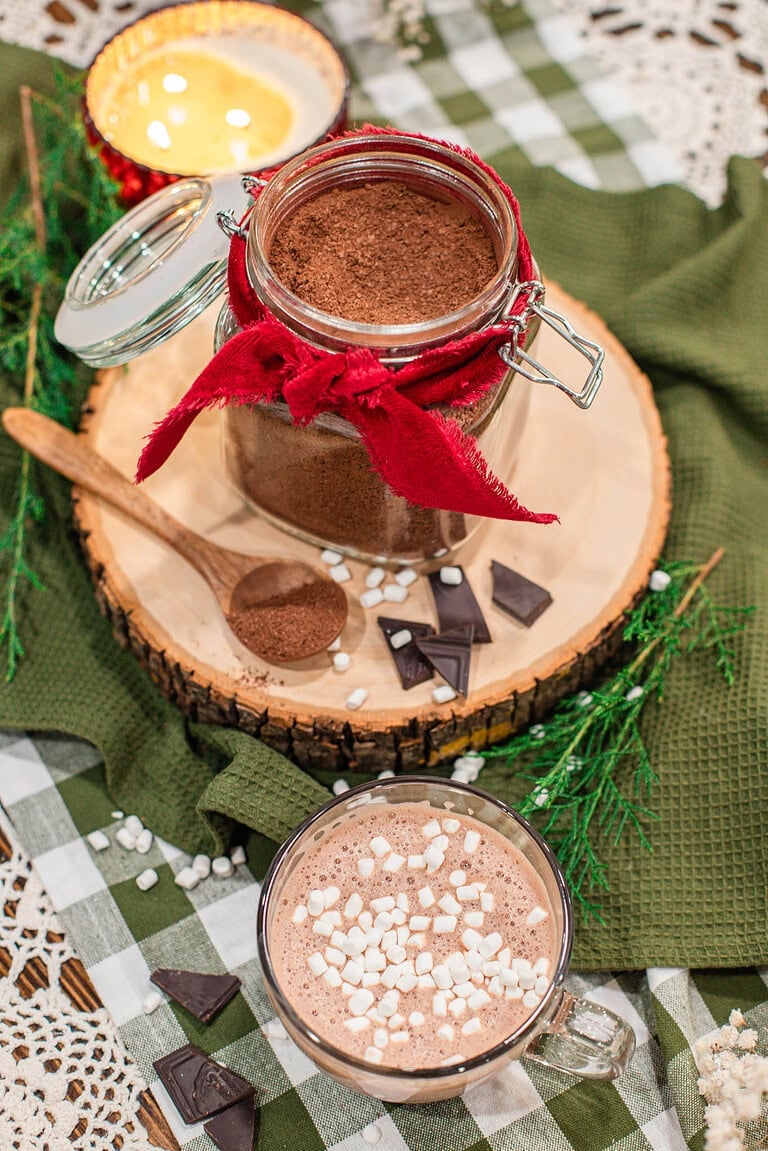 Overhead view of a glass jar of homemade hot cocoa mix tied with a red ribbon on a wood slice, with a wooden spoon of cocoa mix, dark chocolate pieces, and mini marshmallows, beside a glass mug of hot cocoa topped with marshmallows on a green gingham cloth