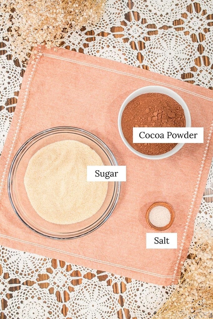 Labeled ingredients flat lay for homemade hot cocoa mix -- a glass bowl of sugar, a white bowl of cocoa powder, and a small wooden bowl of salt -- on a peach linen with dried baby's breath