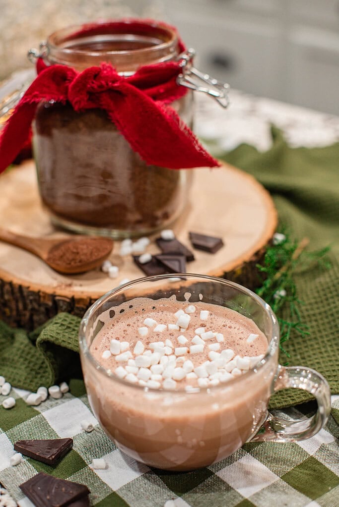 Glass mug of hot cocoa topped with mini marshmallows on a green gingham cloth, with a glass jar of homemade hot cocoa mix tied with a red ribbon, a wooden spoon, and dark chocolate pieces on a wood slice in the background