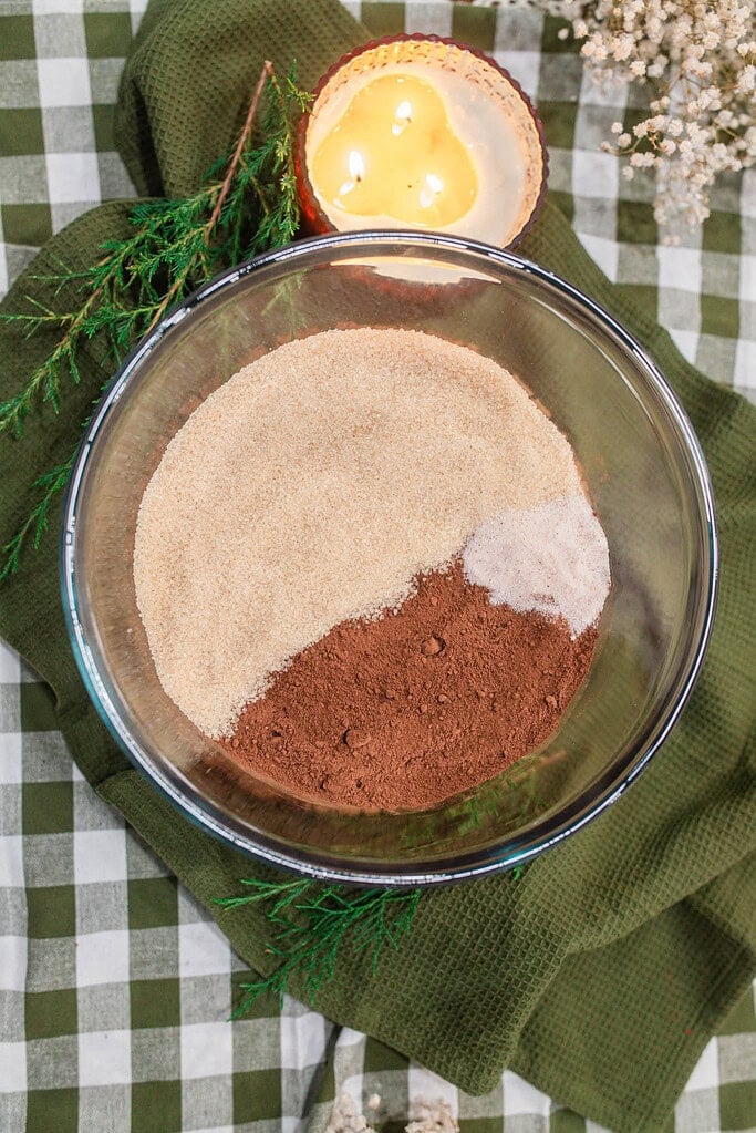 Overhead view of sugar, cocoa powder, and salt measured into a glass mixing bowl before combining into homemade hot cocoa mix, on a green gingham cloth with evergreen sprigs and a lit candle