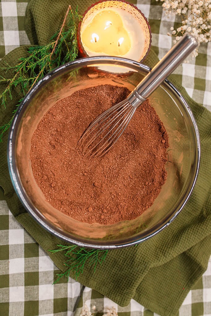 Overhead view of fully whisked homemade hot cocoa mix in a glass bowl with a wire whisk, on a green gingham cloth with evergreen sprigs and a lit candle