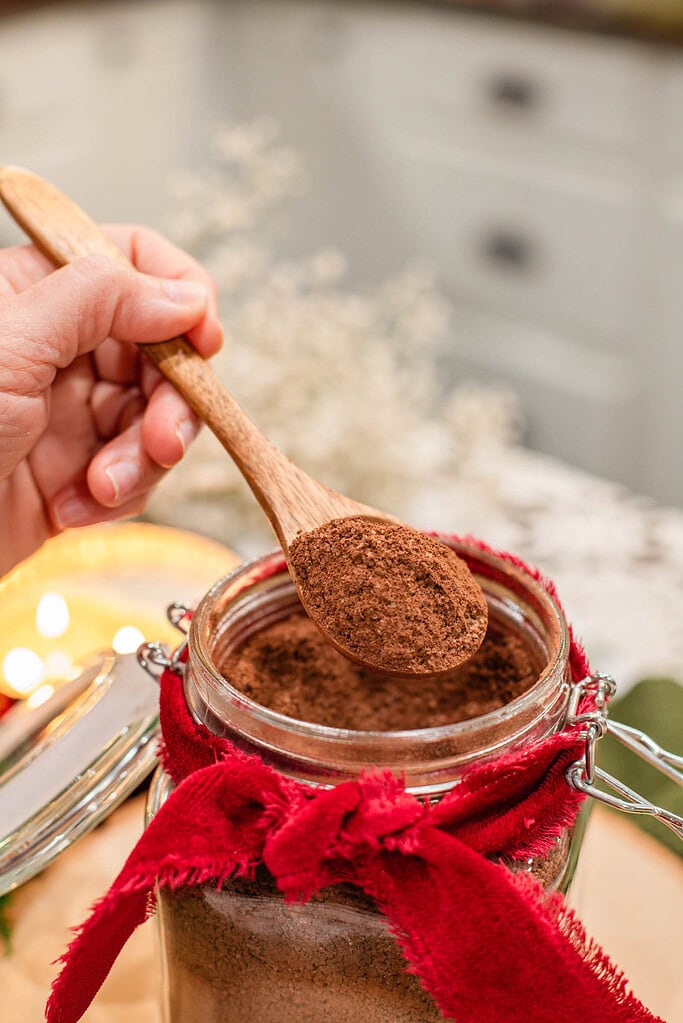 Hand holding a wooden spoon heaped with homemade hot cocoa mix over an open glass jar tied with a red ribbon, with a lit candle and dried flowers in the background