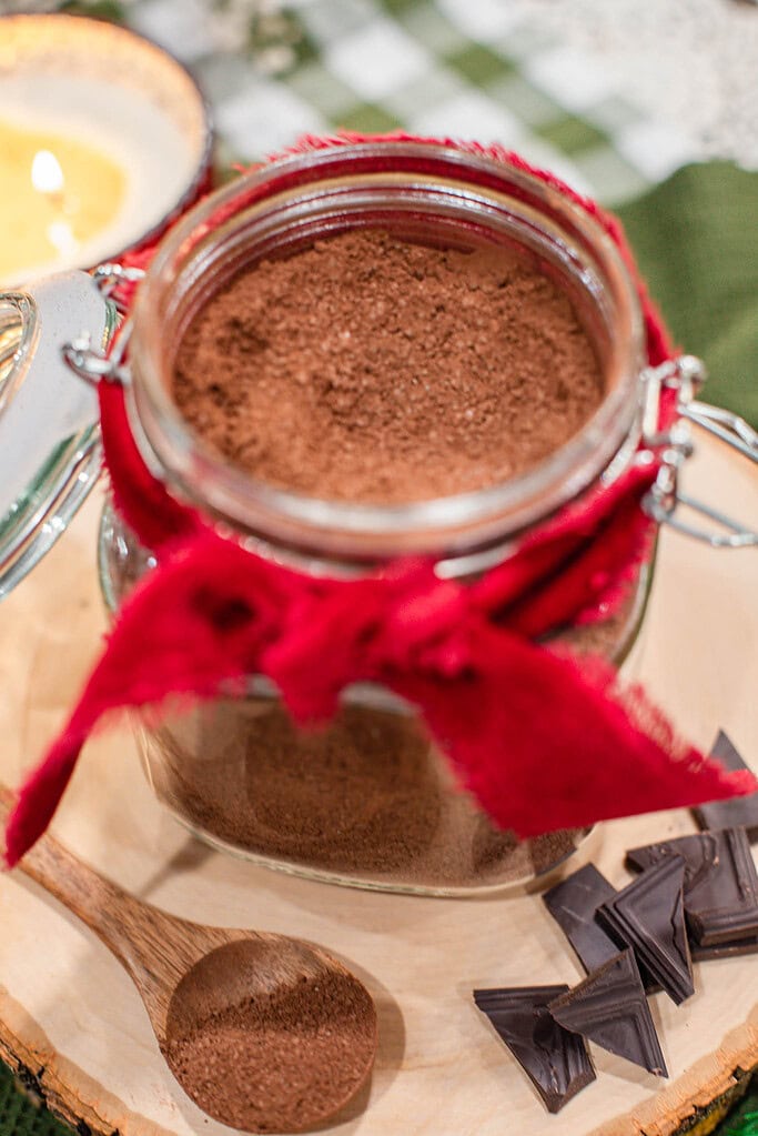 Close-up looking down into an open glass jar of homemade hot cocoa mix tied with a red ribbon, with a wooden spoon and dark chocolate pieces on a wood slice board