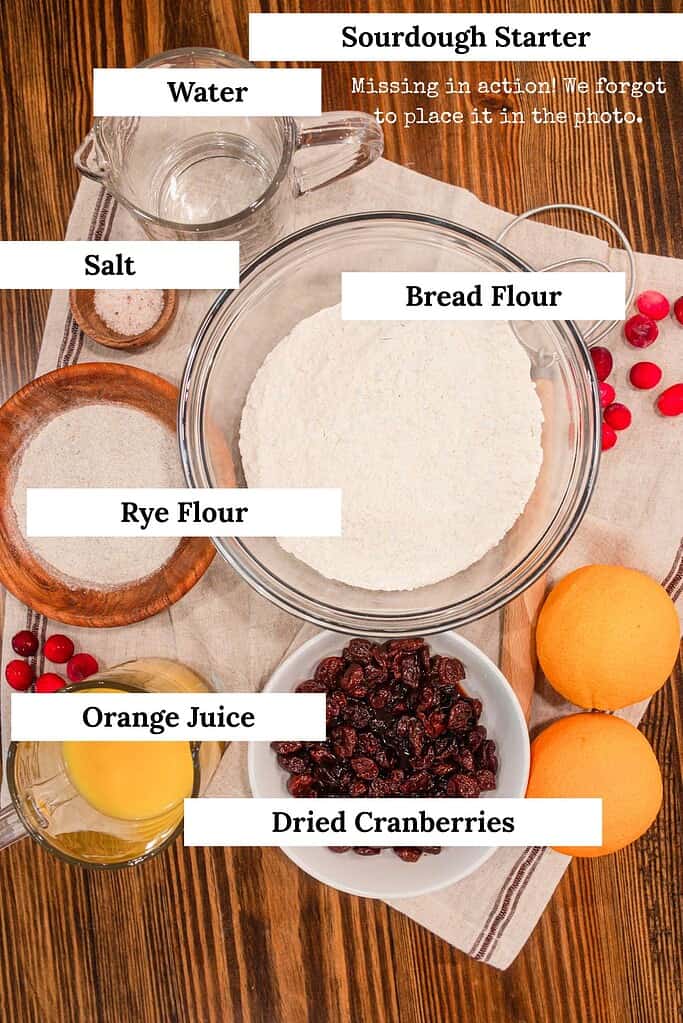 Overhead view of sourdough cranberry orange bread ingredients on a wooden table, including bowls of bread flour, rye flour, salt, dried cranberries, and oranges, with a glass measuring cup of water and orange juice. The sourdough starter is noted as missing from the photo.