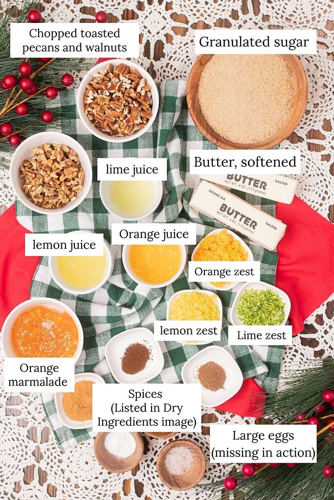 Labeled overhead ingredient flat lay for sourdough fruit cake wet ingredients showing chopped toasted pecans and walnuts, granulated sugar, softened butter sticks, lime juice, lemon juice, orange juice, orange marmalade, orange zest, lemon zest, lime zest, spices, and a note about large eggs being "missing in action" on a green and white buffalo check cloth with pine branches and red ornament berries on crochet lace.