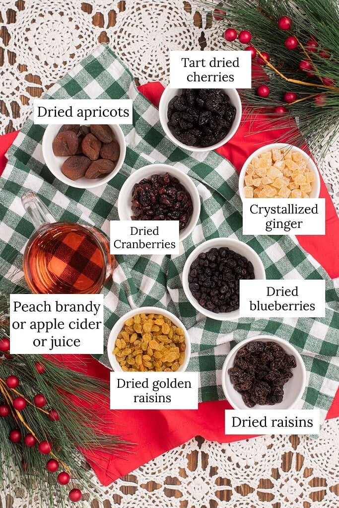 Labeled overhead ingredient flat lay for sourdough fruit cake dried fruit mixture showing dried apricots, tart dried cherries, dried cranberries, crystallized ginger, dried blueberries, dried golden raisins, dried raisins, and peach brandy or apple cider or juice in white bowls and a glass pitcher on a green and white buffalo check cloth with pine branches and red ornament berries on crochet lace.