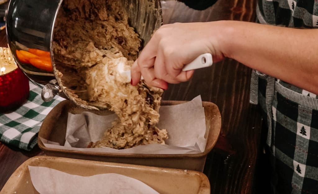 A woman in a kitchen is pouring in uncooked batter into two parchment paper lined loaf pans.