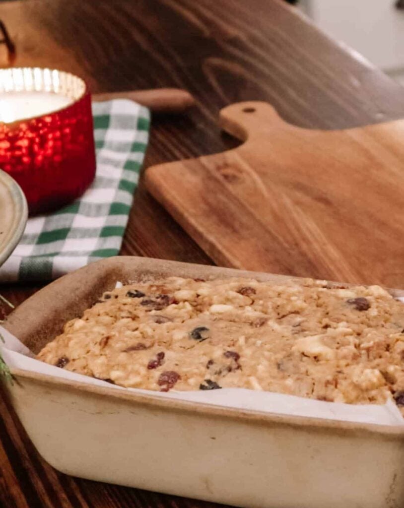 A ceramic loaf pan with fruitcake in it on a wooden counter beside a wooden cutting board and red lit candle in a kitchen.