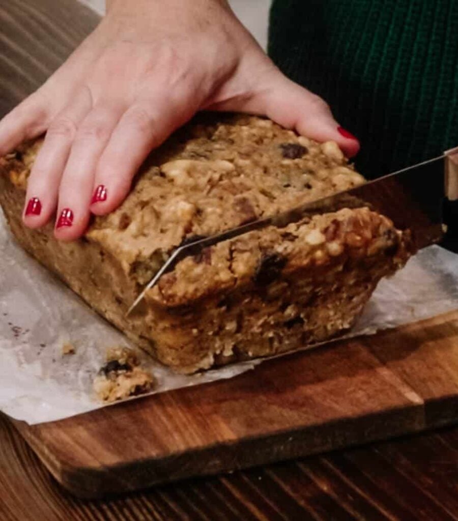 A woman in a kitchen is slicing a sourdough fruitcake on a wooden cutting board.