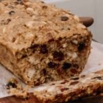 A loaf of freshly baked fruit cake in a cottage kitchen on a wooden counter top on a cutting board with one piece cut off the end.