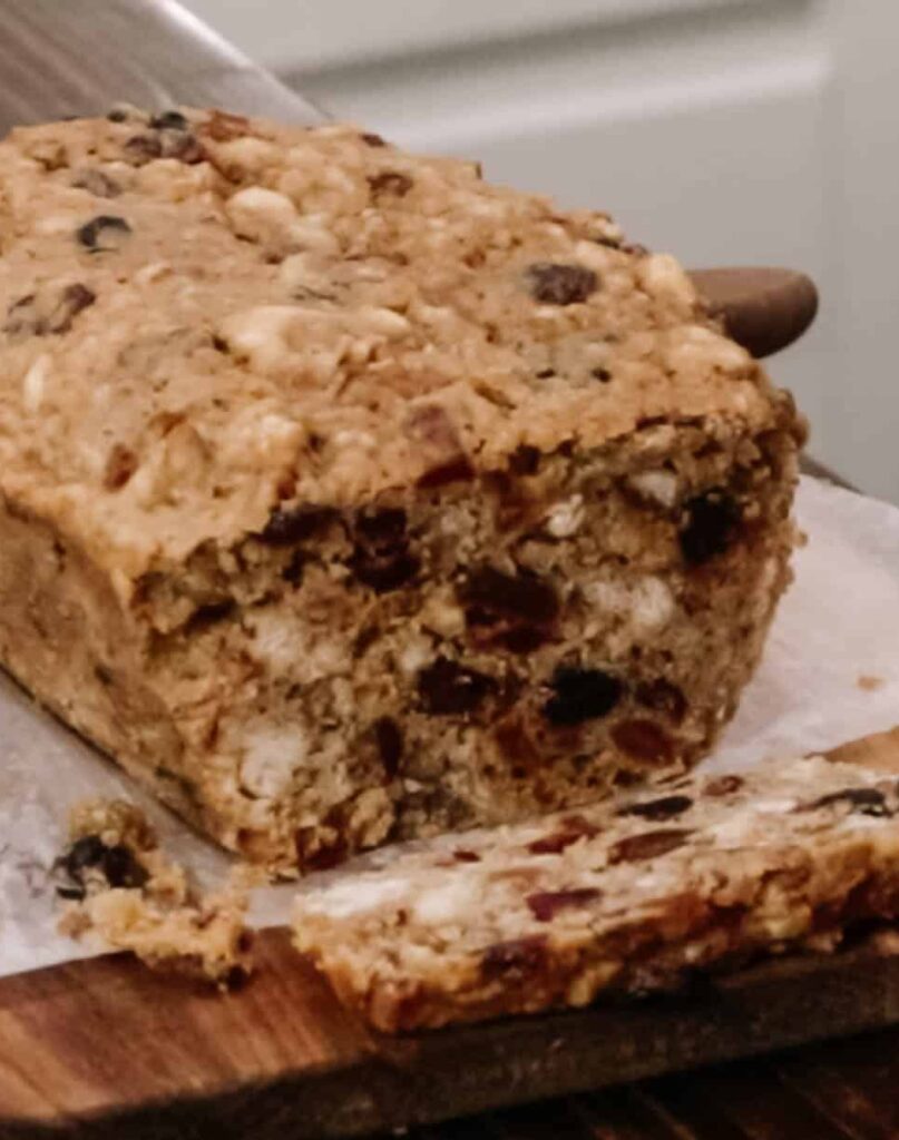 A loaf of freshly baked fruit cake in a cottage kitchen on a wooden counter top on a cutting board with one piece cut off the end.
