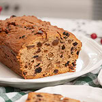 Sliced sourdough fruitcake loaf on a white scalloped plate with one slice plated separately in the foreground, on a green gingham cloth with red cranberries in the background