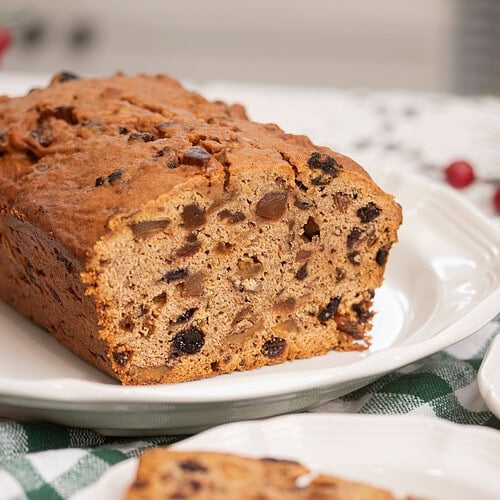Sliced sourdough fruitcake loaf on a white scalloped plate with one slice plated separately in the foreground, on a green gingham cloth with red cranberries in the background