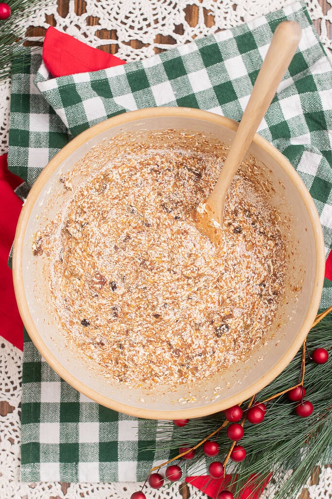 Overhead view of sourdough fruitcake batter with dried fruit and flour being folded together with a wooden spoon in a large mixing bowl, on a green gingham cloth with red berry sprigs