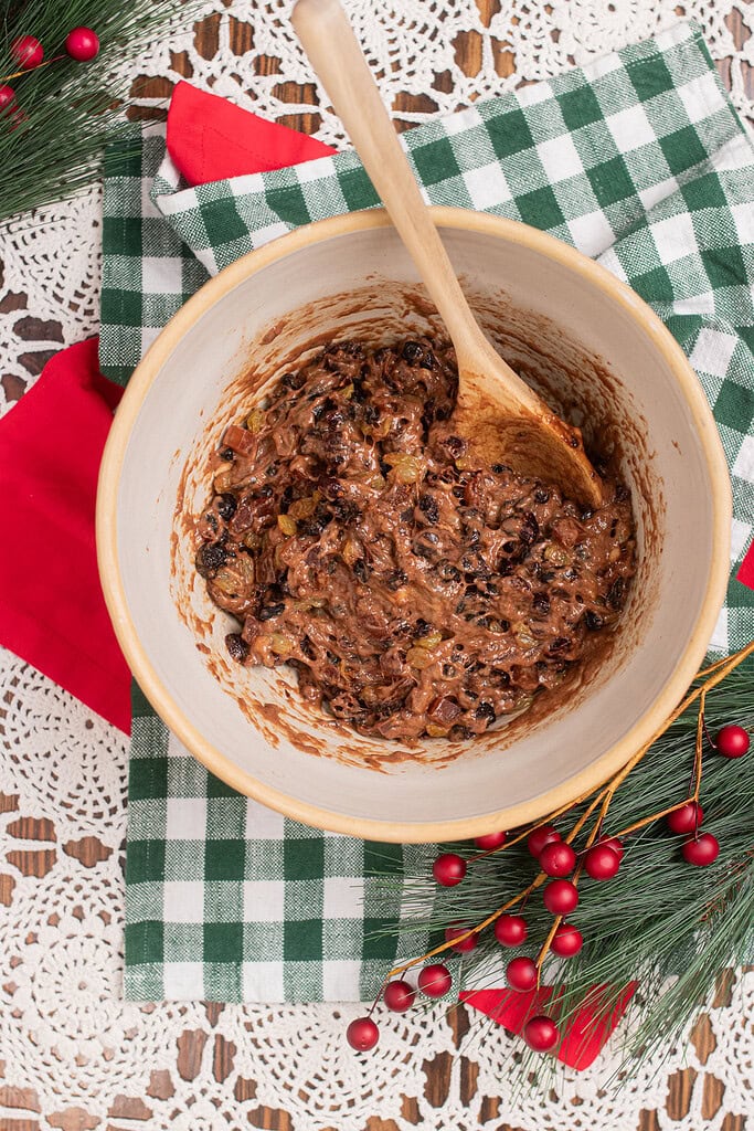 Overhead view of fully mixed sourdough fruitcake batter loaded with dried fruit and raisins in a large mixing bowl with a wooden spoon, on a green gingham cloth with Christmas greenery