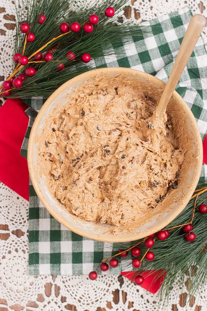 Overhead view of finished sourdough fruitcake batter with dried fruit fully incorporated in a large mixing bowl with a wooden spoon, on a green gingham cloth with pine branches and red berries