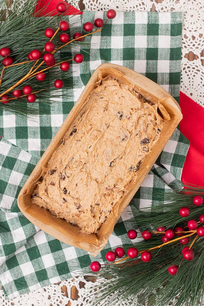 Overhead view of sourdough fruitcake batter spread into a parchment-lined loaf pan ready to bake, on a green gingham cloth with Christmas pine and red berry sprigs