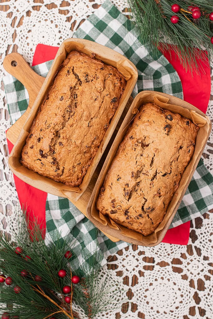 Overhead view of two freshly baked sourdough fruitcake loaves in parchment-lined pans on a wooden cutting board, on a red and green gingham cloth with Christmas greenery and red berries
