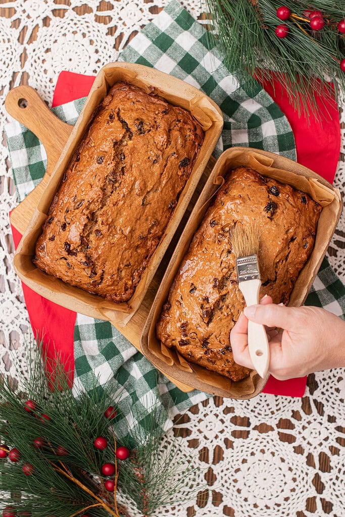 Hand brushing a glaze onto two freshly baked sourdough fruitcake loaves in parchment-lined pans, on a green gingham cloth with pine branches and red berry sprigs