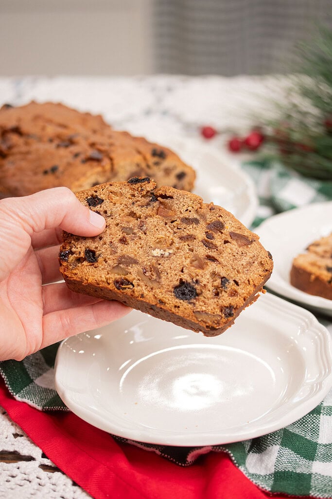 Hand holding a thick slice of sourdough fruitcake studded with dried fruit and nuts over a white plate, with the sliced loaf and Christmas greenery in the soft background