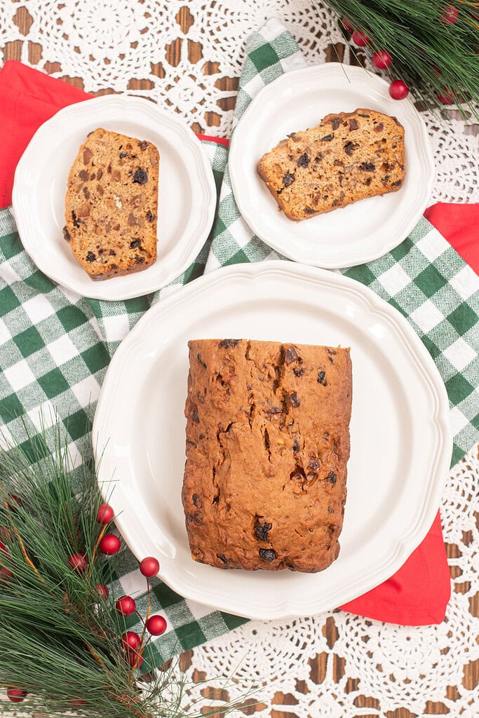 Overhead flat lay of a whole sourdough fruitcake loaf on a large white plate flanked by two smaller plates each holding a slice, on a green gingham cloth with pine branches and red berry sprigs