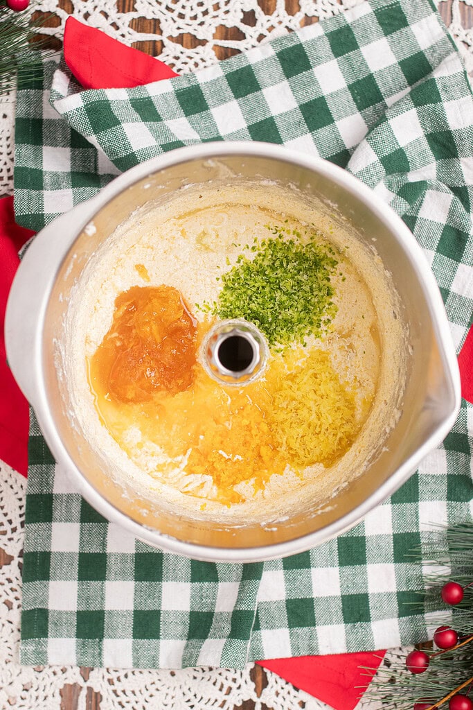Overhead view of a stand mixer bowl with creamed butter and sugar, topped with fresh lime zest, lemon zest, orange zest, and apricot preserves before mixing into sourdough fruitcake batter