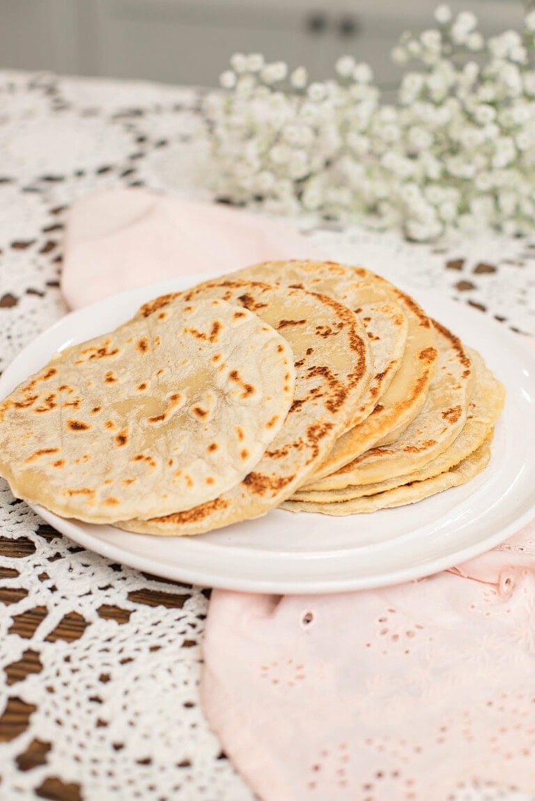 Freshly cooked sourdough tortillas stacked on a white plate, lightly browned with soft bubbles, styled on a lace tablecloth with a blush pink cloth and white flowers in the background.