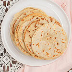 Freshly cooked sourdough tortillas stacked on a white plate, lightly browned with soft bubbles, styled on a lace tablecloth with a blush pink cloth and white flowers in the background.