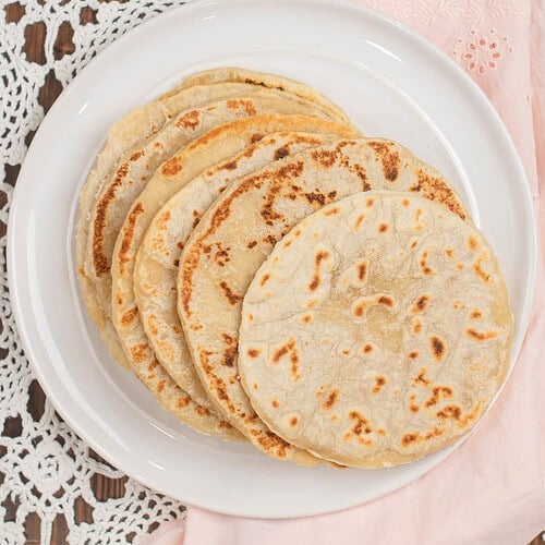 Freshly cooked sourdough tortillas stacked on a white plate, lightly browned with soft bubbles, styled on a lace tablecloth with a blush pink cloth and white flowers in the background.