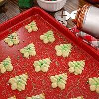 A red baking sheet full of Christmas Tree Spritz Cookies decorated with green sprinkles and gold balls with a vintage cookie press off to the side of the pan.