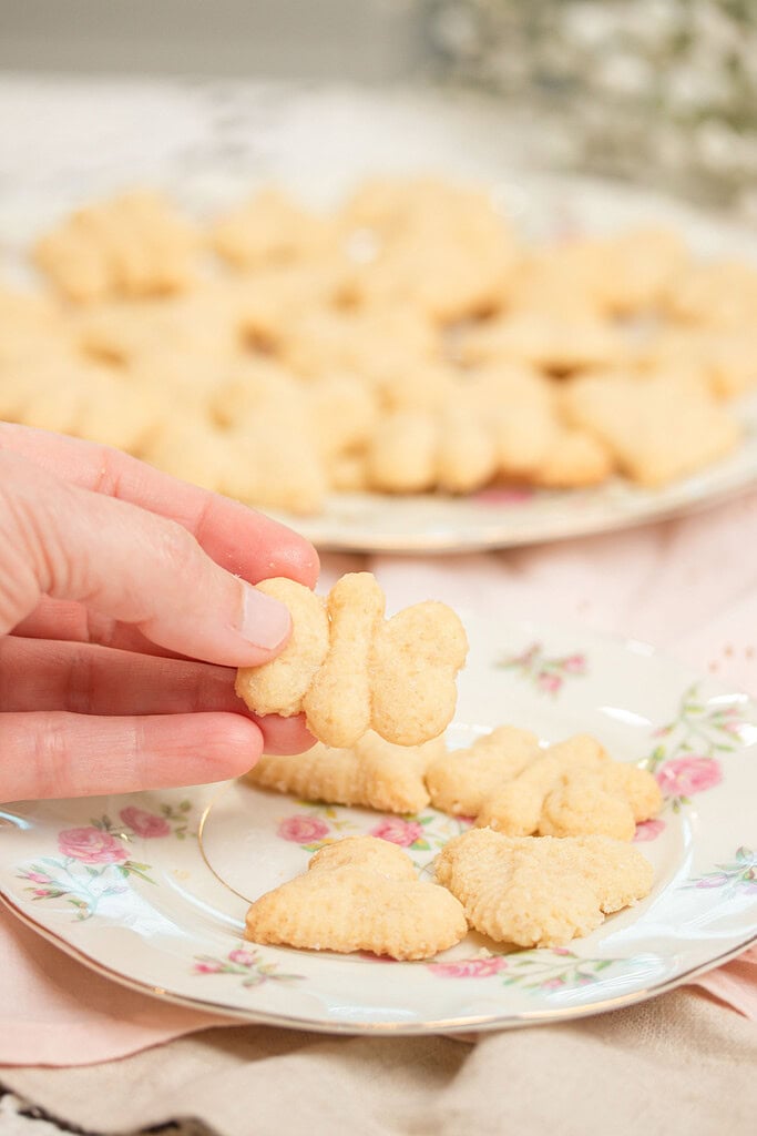 Hand holding a butterfly-shaped spritz cookie over a floral china plate with more spritz cookies, with a full plate of assorted spritz cookies blurred in the background on a pink linen