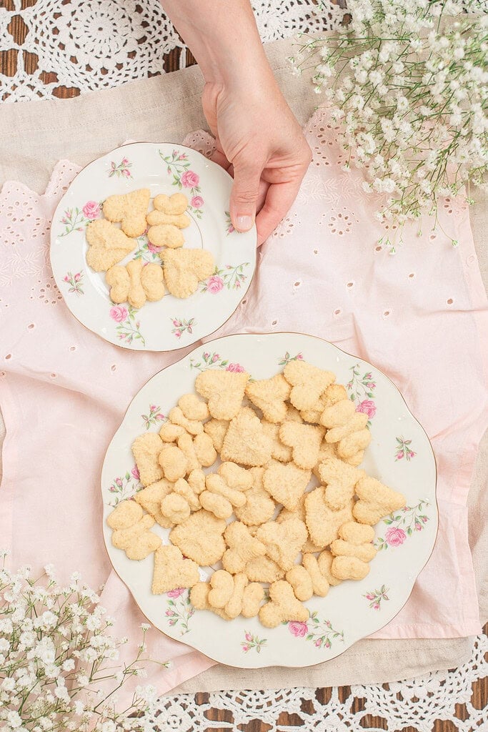 Overhead view of two floral china plates of spritz cookies in assorted shapes on a pink eyelet linen, with a hand reaching to pick up the smaller plate and baby's breath in the corners
