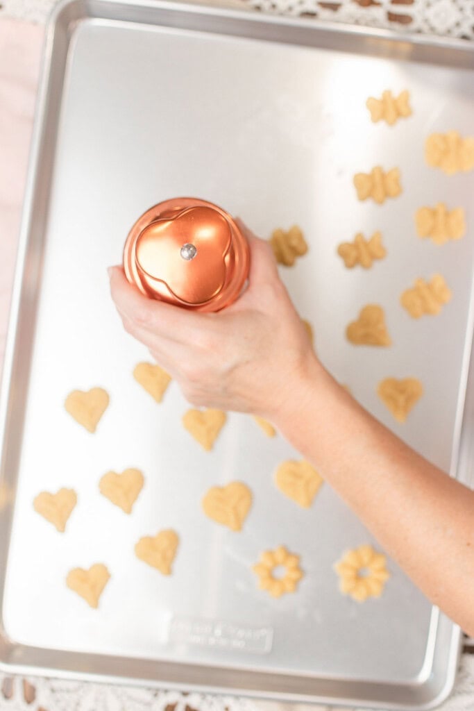 Hand pressing a copper cookie press onto a silver baking sheet already lined with unbaked heart, butterfly, and flower-shaped spritz cookies