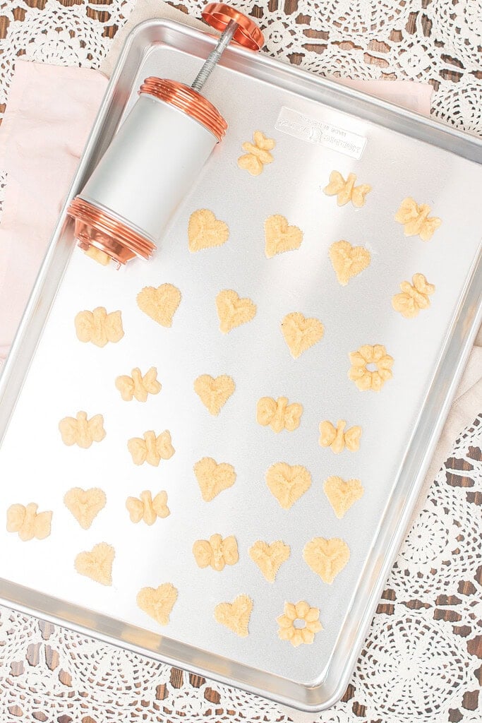 Overhead flat lay of a silver baking sheet filled with unbaked spritz cookies in heart, butterfly, and flower shapes, with a copper cookie press resting in the corner on a lace doily