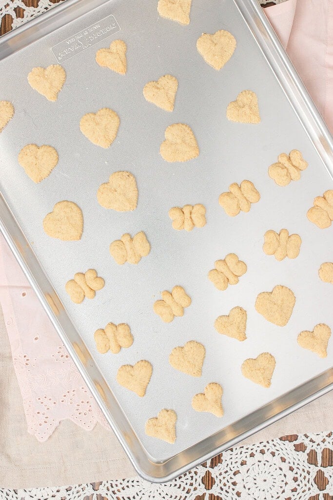 Overhead view of a full silver baking sheet of baked spritz cookies in heart and butterfly shapes on a pink linen and lace doily