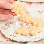 Hand holding a butterfly-shaped spritz cookie over a floral china plate with heart and flower-shaped spritz cookies, with a full plate of spritz cookies blurred in the background on a pink linen