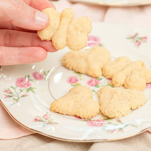 Hand holding a butterfly-shaped spritz cookie over a floral china plate with heart and flower-shaped spritz cookies, with a full plate of spritz cookies blurred in the background on a pink linen