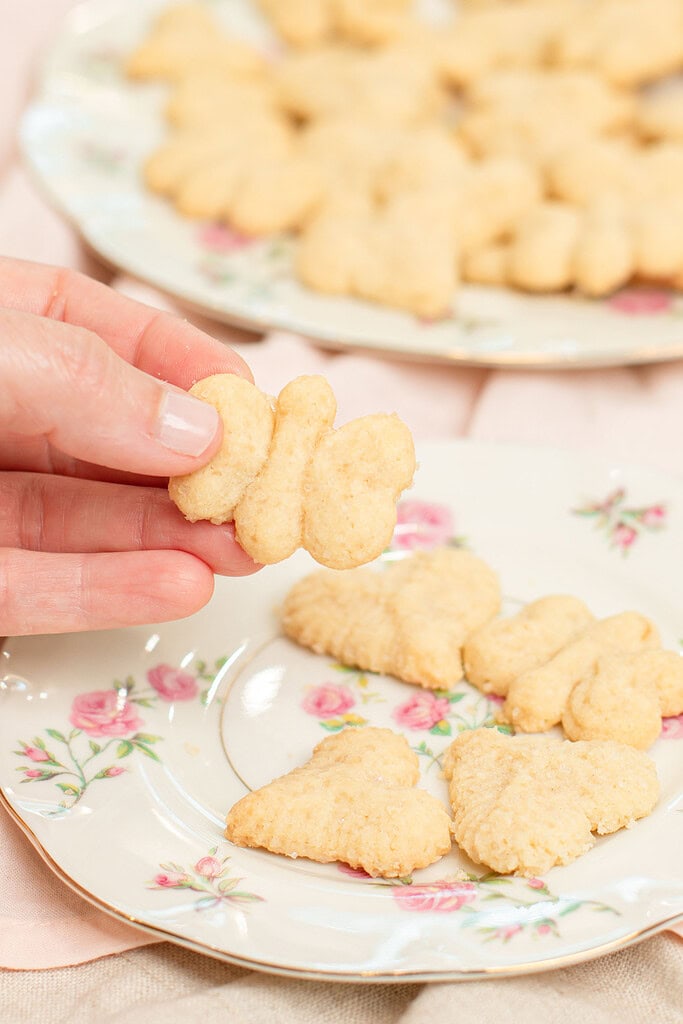 Hand holding a butterfly-shaped spritz cookie over a floral china plate with heart and flower-shaped spritz cookies, with a full plate of spritz cookies blurred in the background on a pink linen