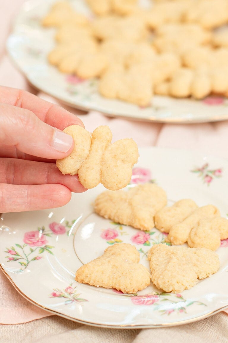 Hand holding a butterfly-shaped spritz cookie over a floral china plate with heart and flower-shaped spritz cookies, with a full plate of spritz cookies blurred in the background on a pink linen
