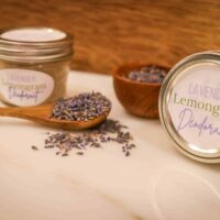 A wooden spoon with lavender buds and two jars of lavender lemongrass deodorant with a small wooden bowl of dried lavender buds on a marble counter top.