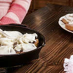 A woman in a pink apron in a kitchen is with another woman in a kitchen also wearing an apron. One woman is icing rolls in a cast iron skillet on a counter top.