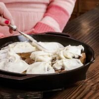 A woman in a pink apron in a kitchen is with another woman in a kitchen also wearing an apron. One woman is icing rolls in a cast iron skillet on a counter top.