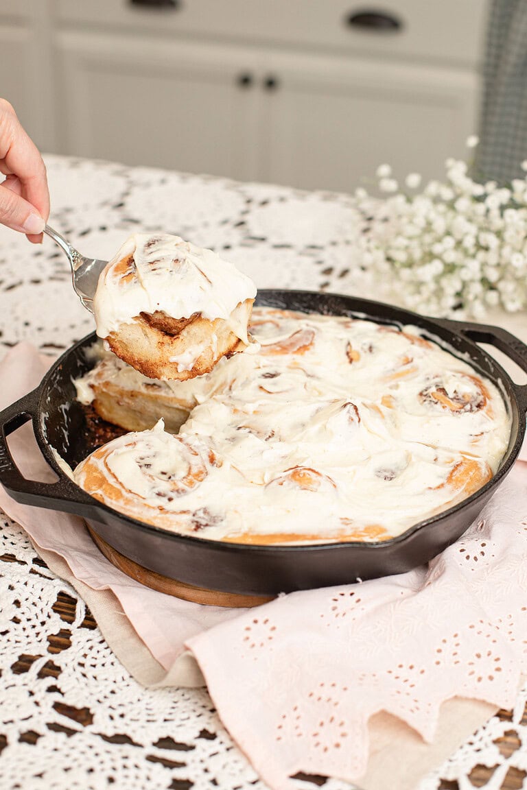 Hand lifting a cream cheese frosted sourdough cinnamon roll from a Lodge cast iron skillet with a spatula, with baby's breath flowers in the soft background on a lace doily