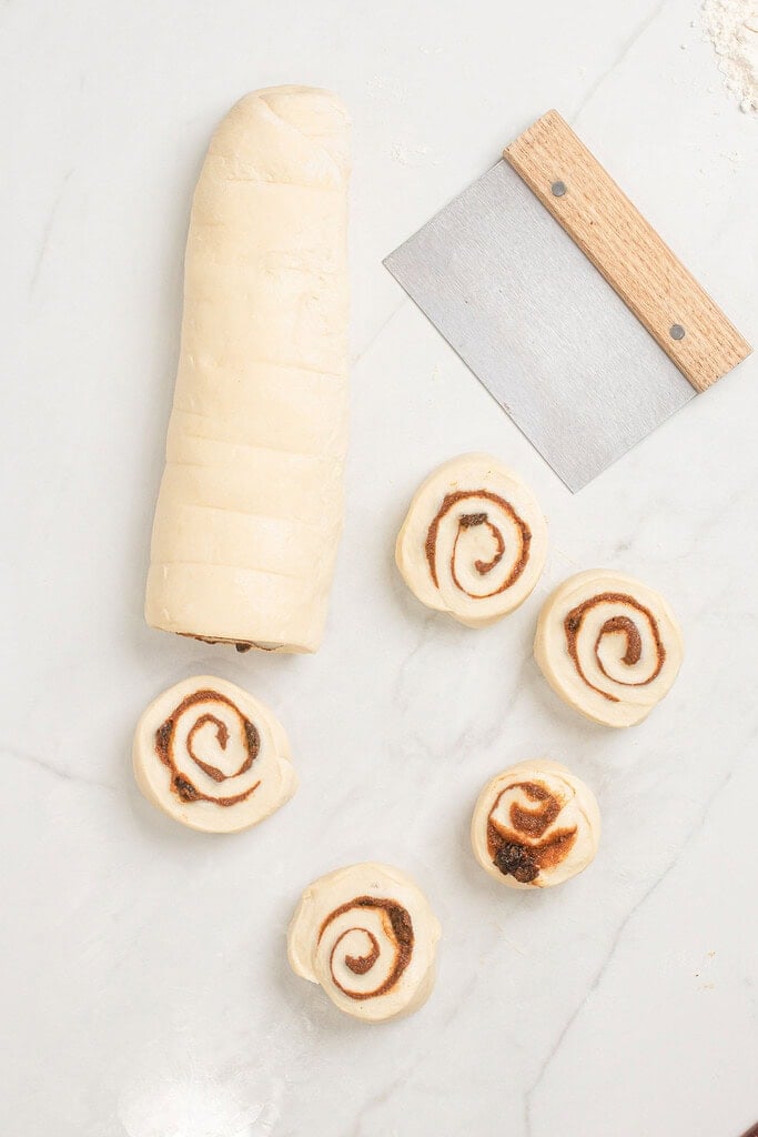 Overhead view of a sourdough cinnamon roll log alongside five individual sliced rolls showing cinnamon swirl cross-sections on a marble surface, with a bench scraper nearby
