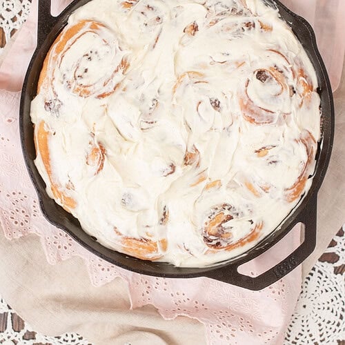 Overhead view of sourdough cinnamon rolls fully covered in cream cheese frosting in a Lodge cast iron skillet on a pink eyelet linen and lace doily