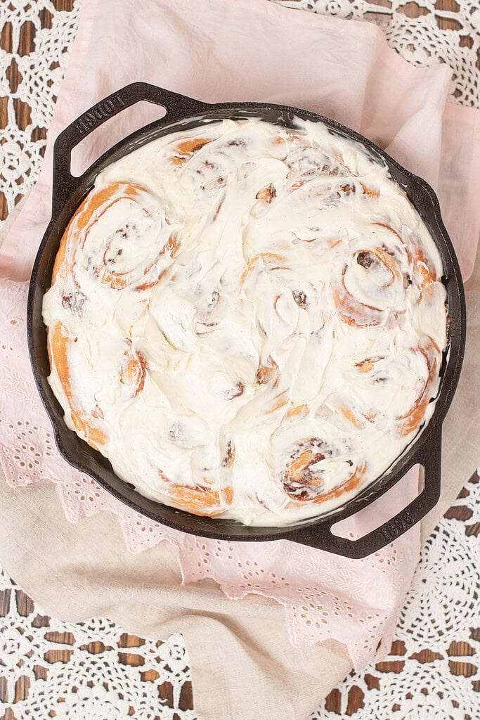 Overhead view of sourdough cinnamon rolls fully covered in cream cheese frosting in a Lodge cast iron skillet on a pink eyelet linen and lace doily