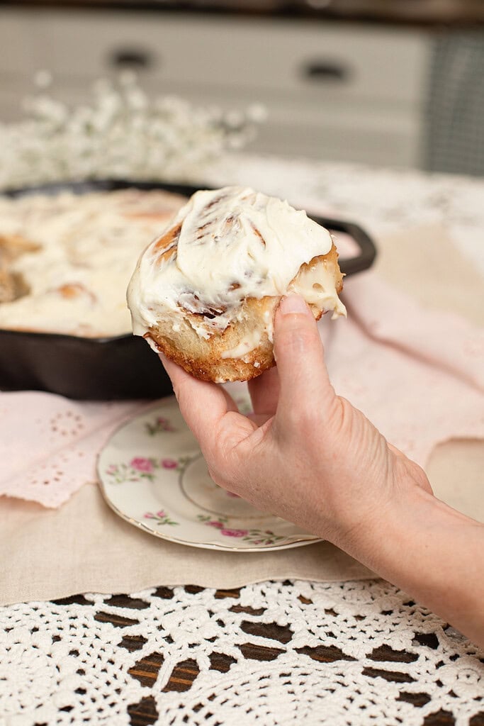 Hand holding a cream cheese frosted sourdough cinnamon roll over a floral china plate, with the cast iron skillet of remaining rolls and baby's breath blurred in the background